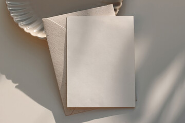 Summer neutral wedding stationery. Closeup of blank greeting card, envelope mockup in sunlight. Invitation, template. Beige dinner plate. Table background, shadows. Elegant composition. Flatlay, top