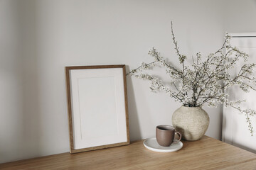 Vertical wooden picture frame, poster mockup in sunlight. Spring, easter composition. Eleganrnterior, home office still life. Blooming cherry plum tree branches in vase. Wooden table. Lateral view.