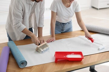 Woman and man applying glue onto wallpaper sheet at wooden table indoors, closeup