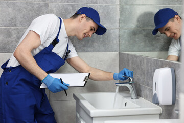 Smiling plumber with clipboard examining faucet in bathroom