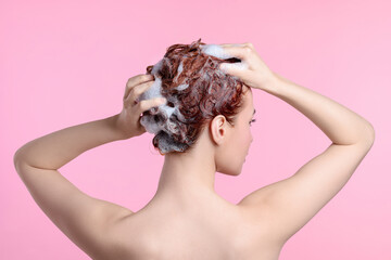 Young woman washing her hair with shampoo on pink background, back view