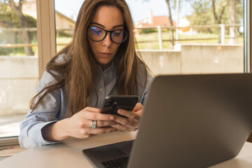 Business woman in light blue shirt and glasses working with her phone and laptop in the office