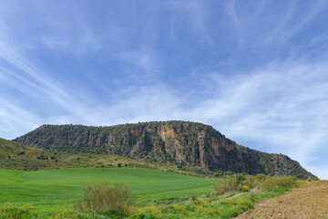 A lush, green meadow stretches out towards a rugged hill, rising prominently beneath a vast, clear...