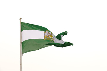 The green and white Andalusian flag, featuring the emblem of Hercules and two lions, is captured as it waves in the breeze under the bright midday sky. There is no other object in the view, highlighti