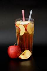 Refreshing fruit juice with ice and straws in a tall glass on a black background, next to red apple slices.
