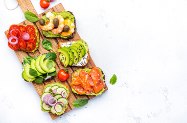 Avocado toasts with salmon, shrimp, vegetables, spinach, capers and cream cheese, served on wooden board, white table background, top view