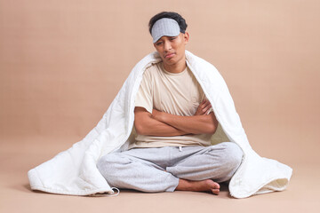 Young man wear eye mask sitting on floor covered with blanket and showing upset expression