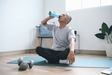 Elderly man drinks water after yoga and sits with crossing legs on exercise mat at home