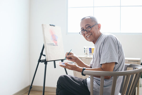 Happy elder man wear glasses sitting on a chair and looking at camera while painting on canvas 
