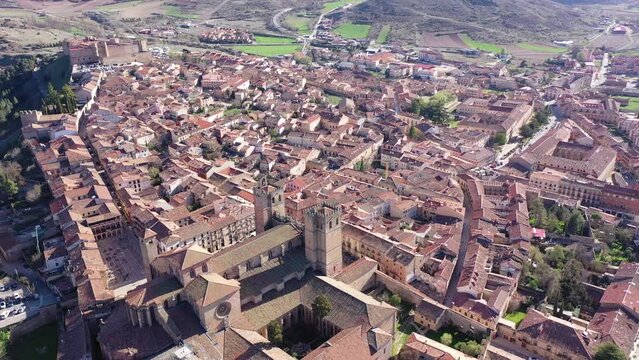General aerial view of Siguenza city with brownish tiled roofs of residential houses, medieval cathedral and fortified castle surrounded by green hills on sunny spring day, Spain
