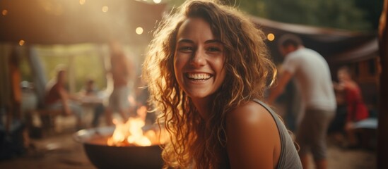A woman with layered hair is smiling happily in front of a fire pit at an outdoor cooking event, enjoying the heat and fun atmosphere by the water - obrazy, fototapety, plakaty