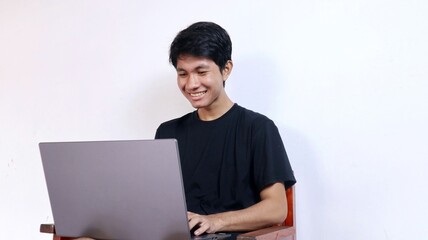 Young happy Asian man with gesture sitting on a chair using a laptop