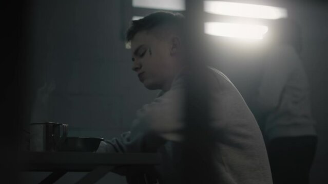 Caucasian teenage prisoner eats prison food at the table in prison cell. Young criminal serves term for crime in youth detention center or jail. Another inmate looks at the window in the background.