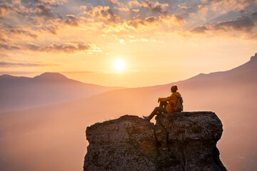 Serene woman hiker is sitting and relaxing on big rock against sunset in mountains