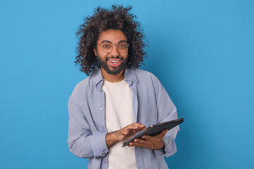 Young cheerful curly Arabian business man with tablet computer in hands looks at screen with smile...