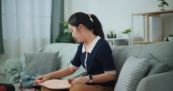 Footage dolly shot, Asian teenager woman sitting on sofa packing travel luggage with clothes for traveling trip, Preparation travel suitcase at home.
