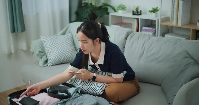 Footage high angle view shot, Asian teenager woman sitting on sofa making checklist of things to pack for travel, Preparation travel suitcase at home.