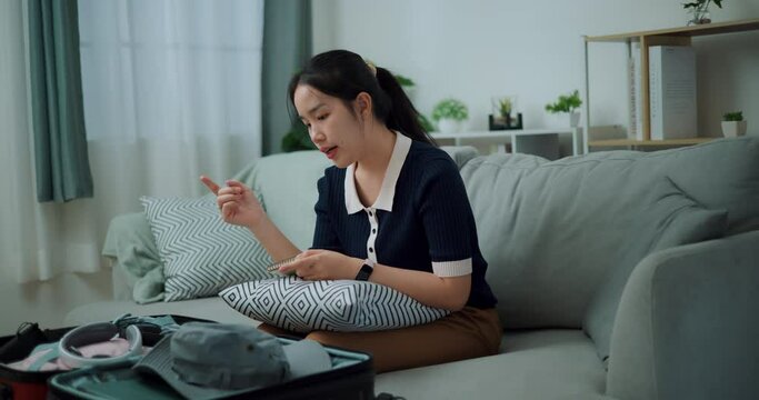 Footage slow motion shot, Asian teenager woman sitting on sofa making checklist of things to pack for travel, Preparation travel suitcase at home.