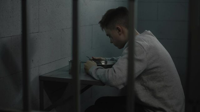 Caucasian guy tries to eat disgusting prison food sitting at the table in prison cell in jail. Teenage criminal, prisoner serves imprisonment term in youth detention center or correctional facility.