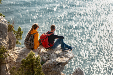 Two young tourists with backpacks are sitting and relaxing on big rock against blue sea