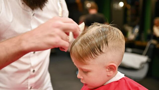 Man Barber Cutting Little Boy's Hair Using Comb And Scissors. Close Up Of Child Getting Haircut From Adult Male, Likely Barber. Professional Hairdresser And Cute Client At Modern Barbershop.