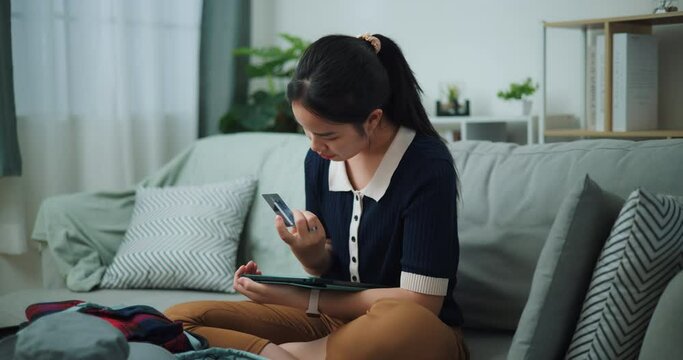 Footage slow motion shot, Asian teenager woman sitting on sofa holding credit card making online payment on digital tablet, Preparation travel suitcase at home.