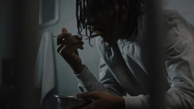 Close up of African American teenage criminal trying to eat disgusting prison food sitting on bed in prison cell. Young prisoner serves imprisonment term for crime in jail. Juvenile detention center.