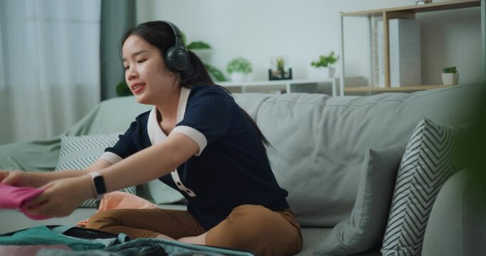 Footage dolly shot, Asian teenager woman wear wireless headphones sitting on sofa packing travel luggage with clothes for traveling trip, Preparation travel suitcase at home.