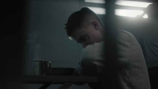 Caucasian teenage criminal eats prison food at the table in prison cell in jail. Young prisoner serves imprisonment term in juvenile detention center. Another inmate looks at window in the background.
