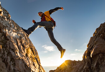 Young sporty hiker with backpack jumps to big rock against sunset sky. Active lifestyle concept