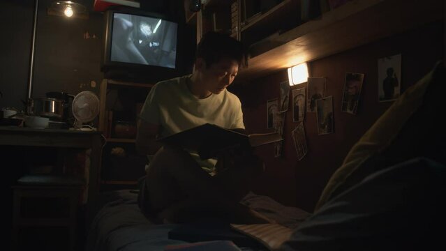 Full footage of young Chinese woman sitting on bunk bed in dark tiny capsule flat, reading textbook, getting ready for college exam, and TV set playing horror movie in backgroundFull footage of young 