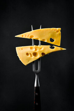 Two pieces of Maasdam cheese on a metal fork. On a black background, vertical photo.
