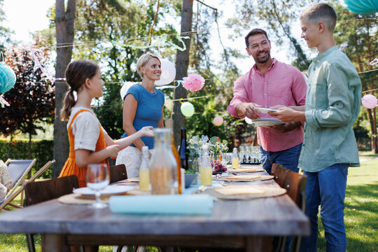 Whole family setting table for summer garden party. Bringing plates, food, and drinks.