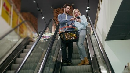 Lovely family couple standing on escalator inside shopping area. Attractive marriage people browsing social medias of modern smartphone and holding shopping basket with various items together.