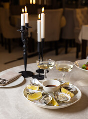 open oysters on plate with slice of lemon on ice cubes on table.candles in the chandelier in restaurant, woman hand taking dressing with spoon putting in sea fruit.romantic dinner