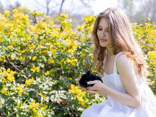 Naklejka na ściany i meble beautiful delicate woman with black bunny rabbit in palms against blossom tree flowers or yellow on bush. smiling blonde girl in white dress in park or garden, sunny spring day. easter is coming.