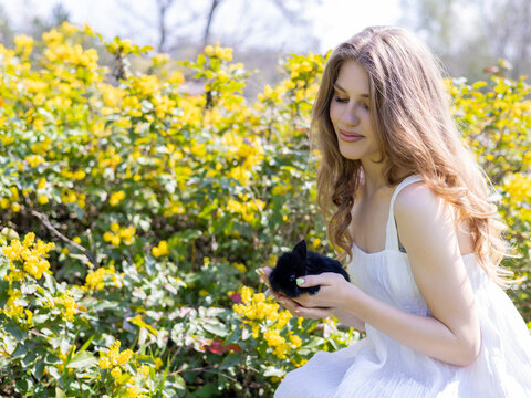 beautiful delicate woman with black bunny rabbit in palms against blossom tree flowers or yellow on bush. smiling blonde girl in white dress in park or garden, sunny spring day. easter is coming.
