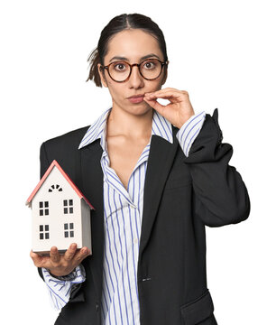 Caucasian businesswoman with house model, real estate concept on studio background with fingers on lips keeping a secret.