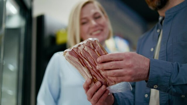 Bearded caucasian husband holding packaged stack of bacon and discussing it lovely wife in supermarket. Friendly family reading product composition and talking about dish that preparing from it.