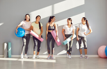 Fototapeta na wymiar Group of young sporty girls, attractive slim happy people posing in fitness club, sportswomen team holding yoga mats. Active friends exercise or sport instruction, recreational activity together 