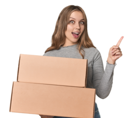 Blonde woman with cardboard boxes moving on studio backdrop smiling and pointing aside, showing something at blank space.