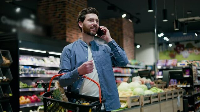 Handsome caucasian male standing in grocery store holding shopping basket and engaged in modern smartphone conversation. Young bearded man solving work issues online while buying food products.