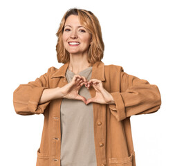 Blonde middle-aged Caucasian woman in studio smiling and showing a heart shape with hands.