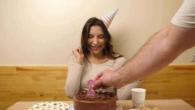 A girl sits in front of a table with a festive cake, into which a candle in the form of the number 22 is stuck. The concept of a birthday celebration.