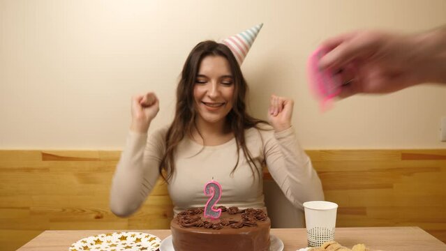 A girl sits in front of a table with a festive cake, into which a candle in the form of the number 26 is stuck. The concept of a birthday celebration.