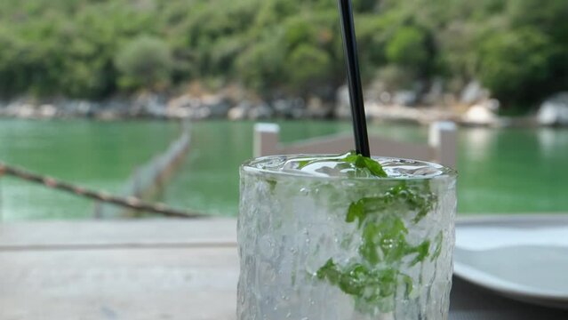 Super slow motion macro of falling fresh aromatic leaf of peppermint herb falling in transparent glass with soda and ice cubes during mojito cocktail preparation by bartender in bar or restaurant. 4k 