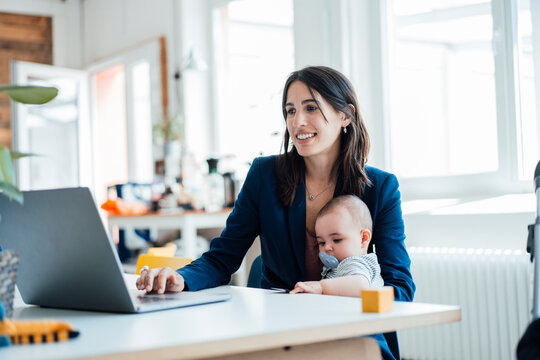 Smiling businesswoman sitting with baby girl using laptop at home