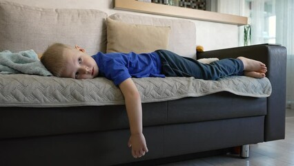 A lazy, bored child is lying on the couch at home. The blond boy wastes time watching TV uselessly. Children's dependence on gadgets. 4K ultra HD quality video footage of a real people domestic life