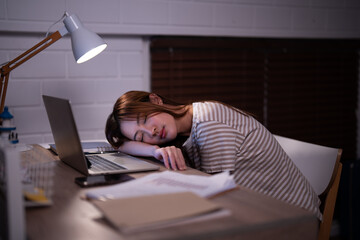 Asian student girl overtaken by fatigue has fallen asleep on their study material after created presentation with the light of a desk lamp illuminated in bedroom at the bedtime - obrazy, fototapety, plakaty