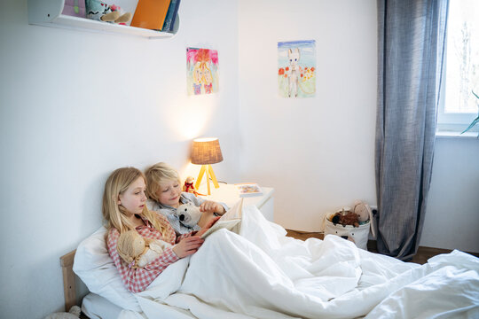 Blond brother and sister reading book on bed at home